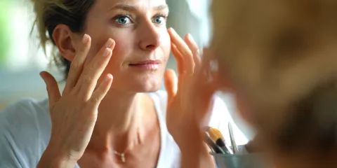 A woman looks in the mirror and carefully applies a skincare product to her cheekbones.