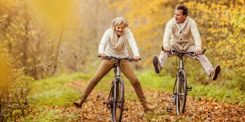 An older man and woman riding bikes through fall foliage with their feet raised playfully off the pedals.