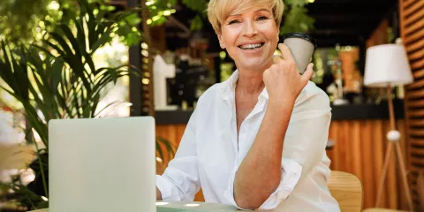 Professional woman sits outdoors working at her laptop with a coffee and smiles into the camera.