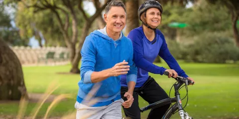 Couple in 50's in park, man with blue sweatshirt running & woman in blue shirt on her bike