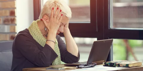 Middle aged woman at desk with laptop, stressed with hands on her head