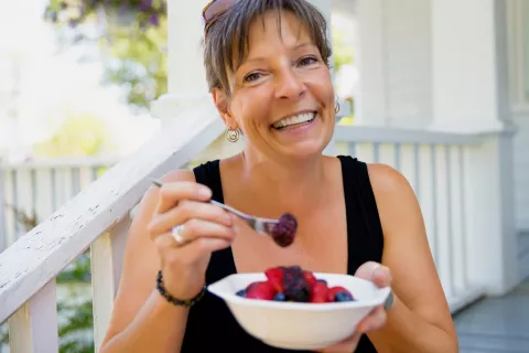 Woman smiling, eating a bowl of colorful fruit on front porch steps 