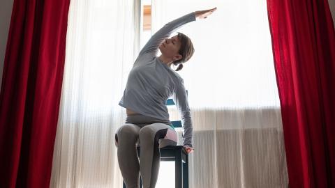 Woman in grey workout clothes stretches her side while seated in a chair