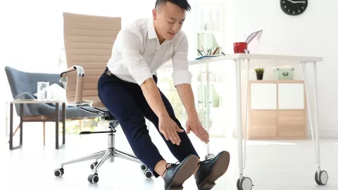 Man sits at the edge of his office chair and reaches his hands out to touch his toes with legs extended