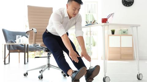 Man sits at the edge of his office chair and reaches his hands out to touch his toes with legs extended